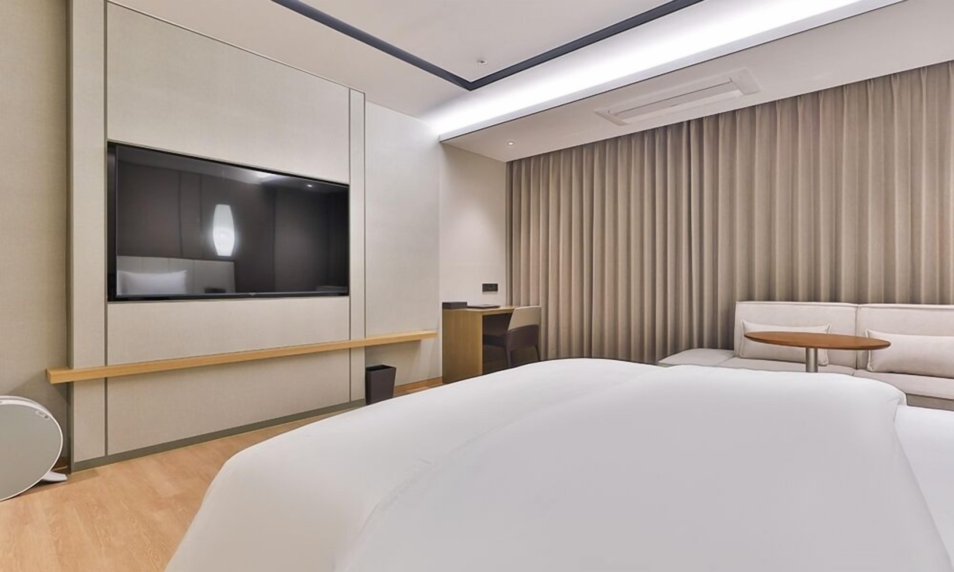 A modern hotel room with a large bed, a flat-screen TV mounted on the wall, a desk with a chair, and a small round table.