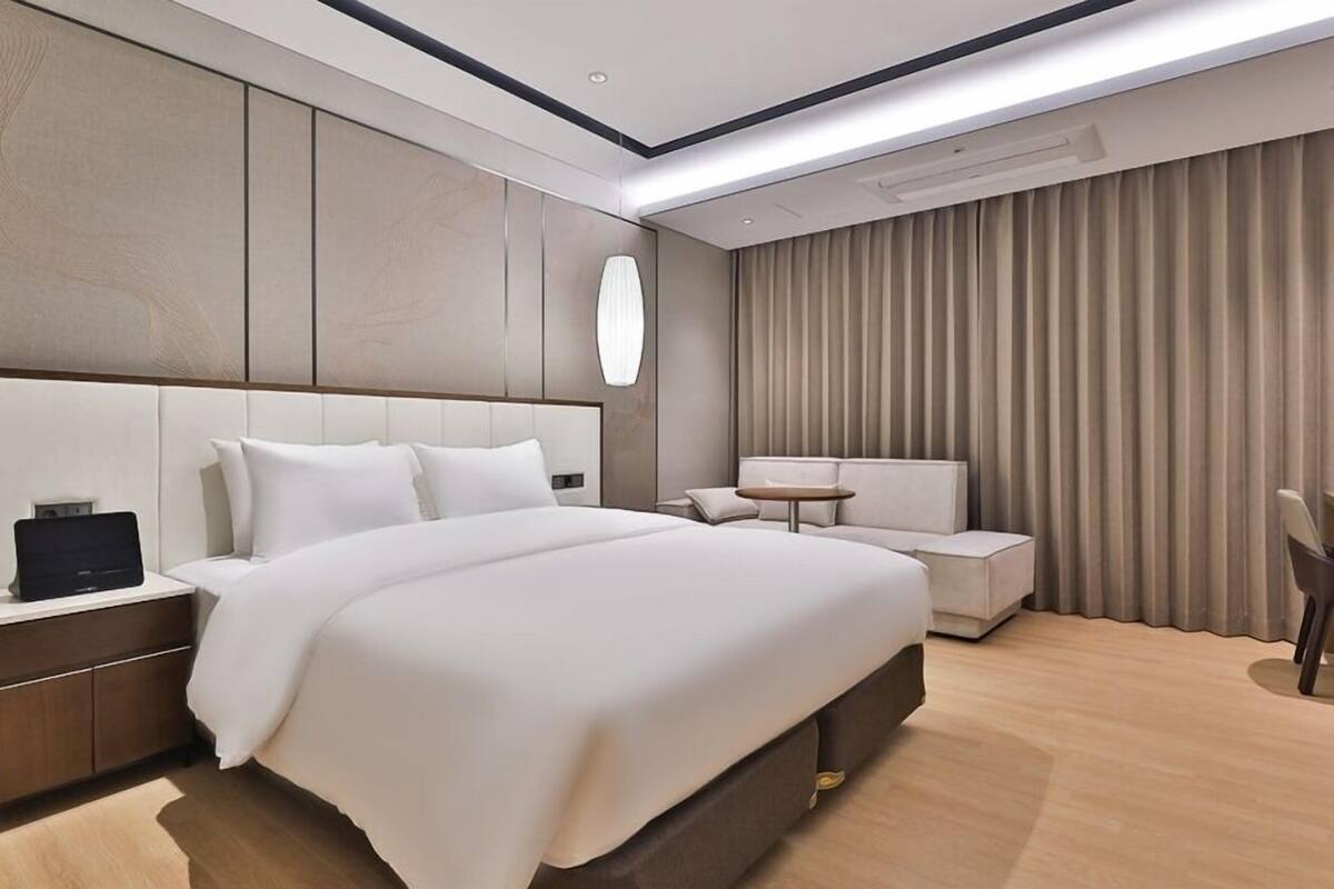 Une chambre d’hôtel moderne dotée d’un grand lit, d’une table de chevet avec une lampe, d’un coin salon avec une petite table et d’une grande fenêtre avec des rideaux.