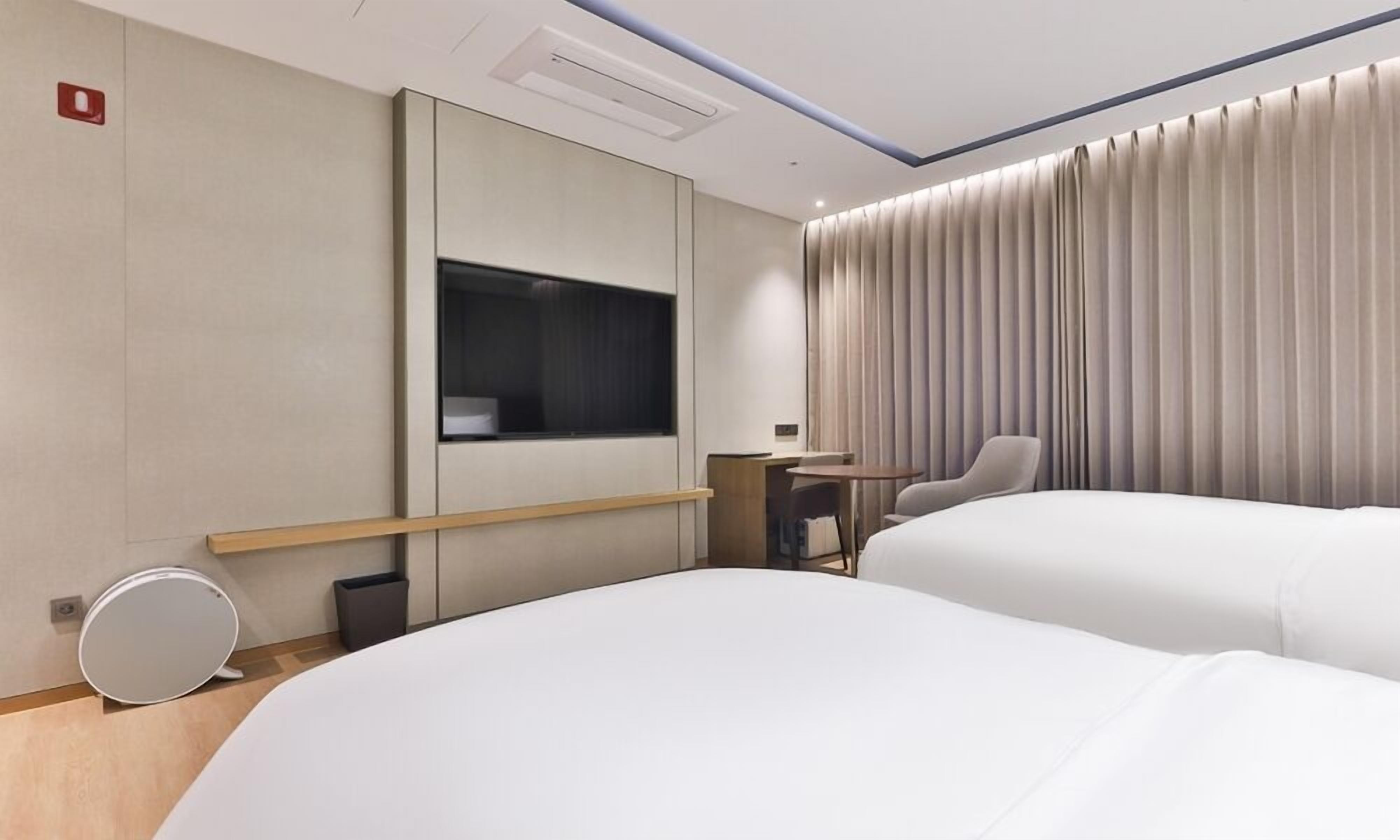 A modern hotel room with two beds, a desk, a chair, and a large window with curtains.
