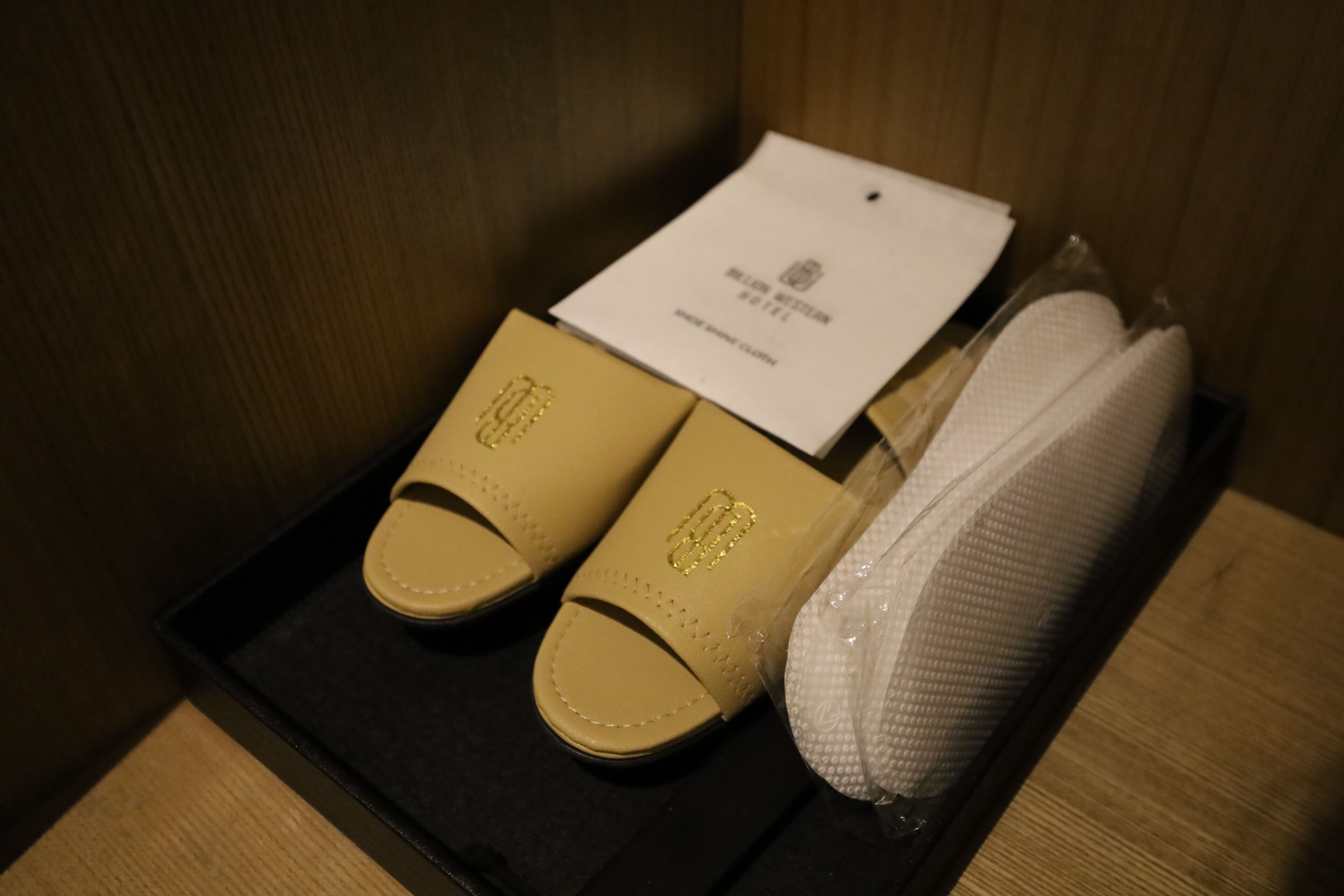 Un par de zapatillas beige con un emblema dorado, un pañuelo blanco y una caja negra con una etiqueta.