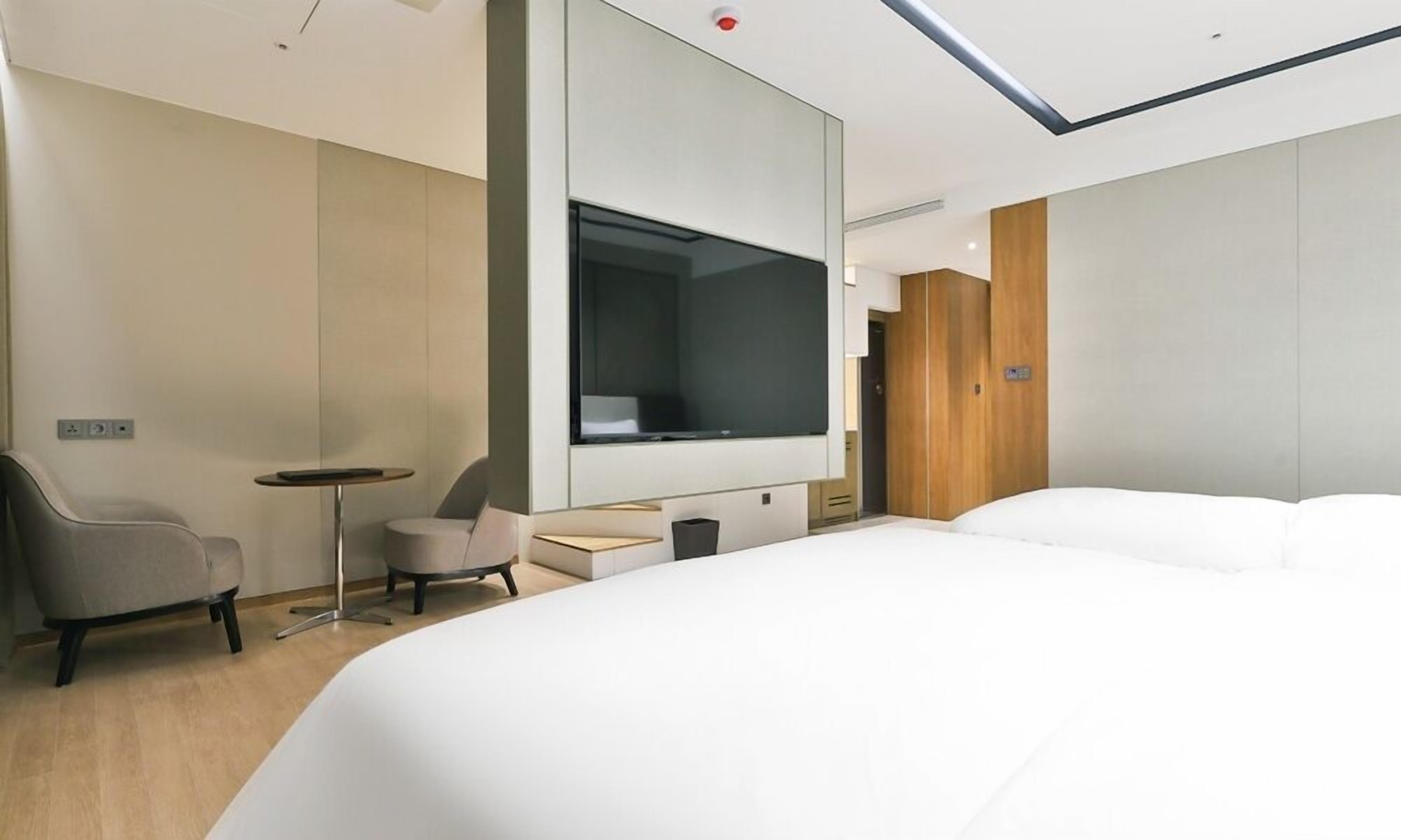 A modern hotel room with a large bed, a small desk, a chair, a TV, and a wardrobe.