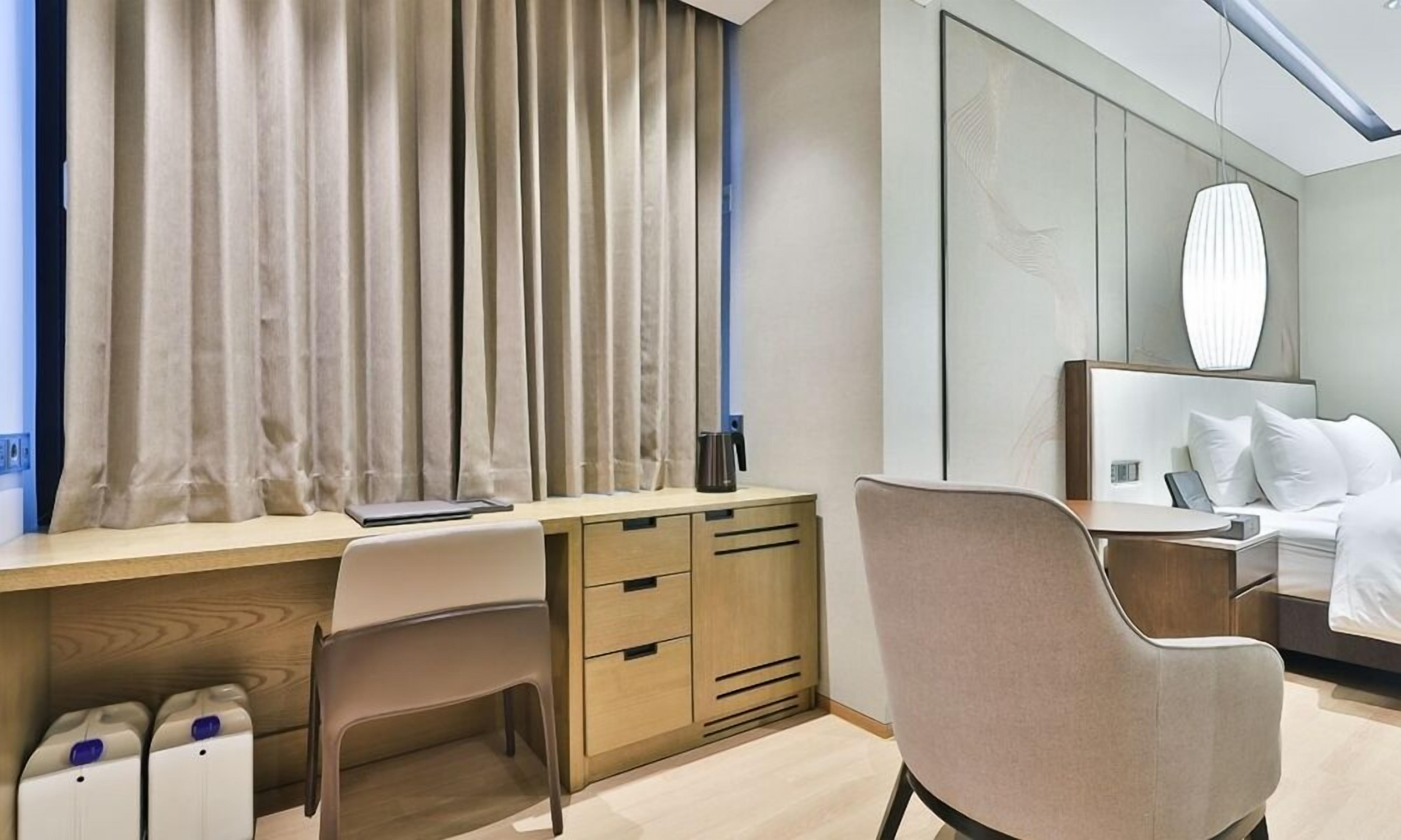 A modern hotel room with a wooden desk, a comfortable chair, a bed with white bedding, and a large mirror above the bed.