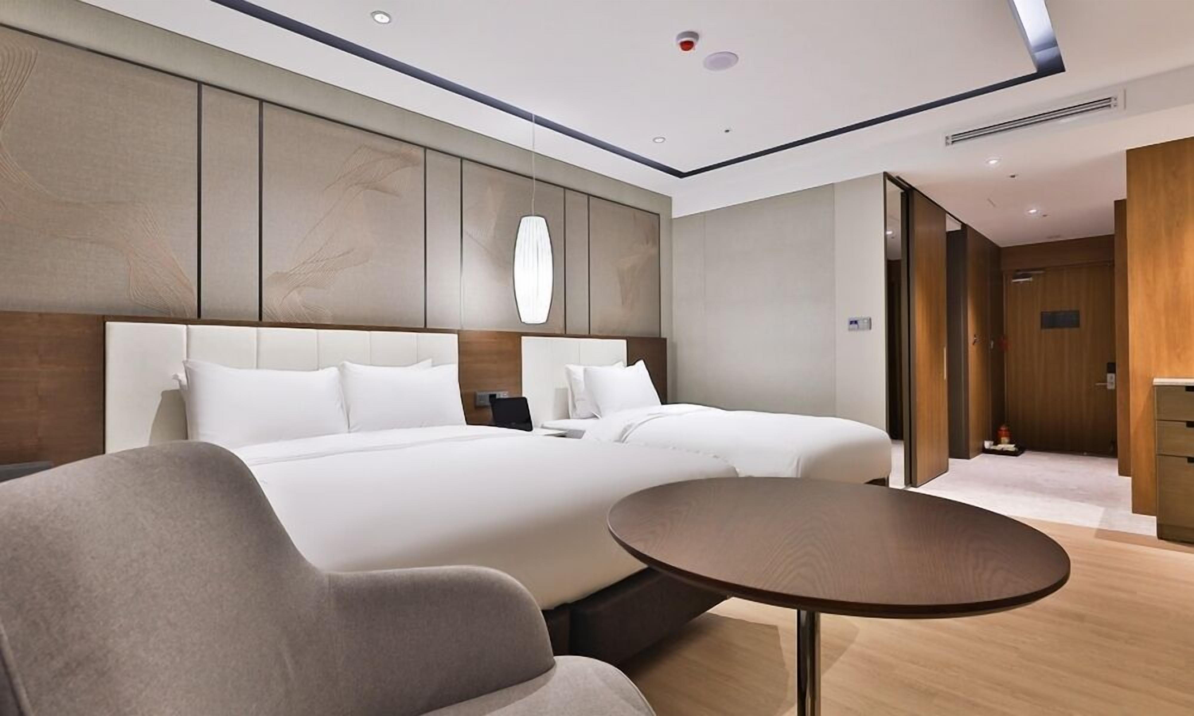 Una habitación de hotel moderna con una cama grande, una mesita de noche, una mesa de centro redonda y vistas al pasillo.