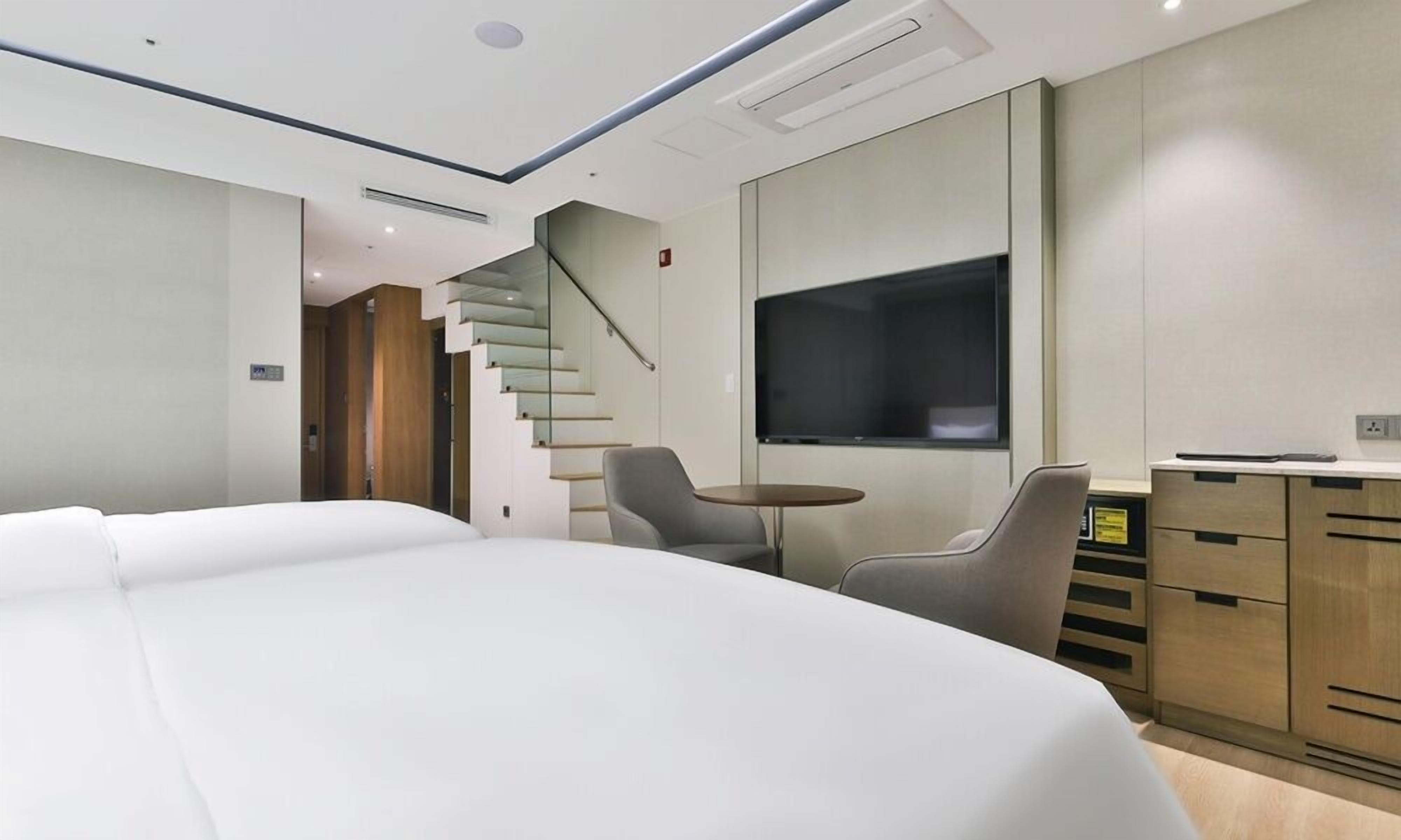 A modern hotel room with a large bed, a TV, a desk with drawers, and a staircase.