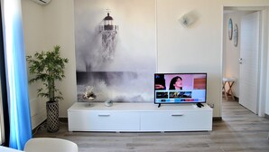 A modern living room with a large flat-screen TV on a white entertainment unit, a potted plant, and a wall-mounted artwork depicting a lighthouse. - Dinky Home Air-conditioned penthouse with sea view terrace (Formia)