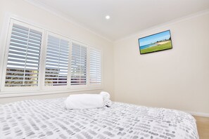 A bedroom with a bed, white blinds, a wall-mounted TV, and a beach view. - Point Bulli (Bulli)