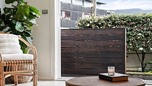 A wicker chair with a cushion, a round wooden table with a book and a candle, and a wooden partition with a view of a garden and mountains in the background. - Point Bulli (Bulli)