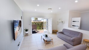 A modern living room with a grey sofa, a coffee table, and a TV mounted on the wall. There is a sliding glass door leading to an outdoor area with a wooden deck and a garden. - Point Bulli (Bulli)