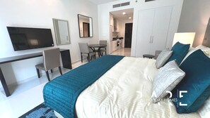 A hotel room with a bed, a desk, a chair, a TV, a mirror, and a kitchenette.