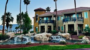 Un hotel de varios pisos con balcones, un jardín ajardinado con una cascada y palmeras.