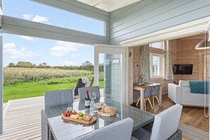 A spacious outdoor dining area with a glass table, surrounded by chairs, and a view of a green field. - Hayditch, Great Field Lodges -  a lodge that sleeps 2 guests  in 1 bedroom (Braunton)