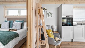 Een gezellige slaapkamer met een bed, nachtkastje en een houten ladder. Een keuken met kastjes en een Bosch oven is zichtbaar.