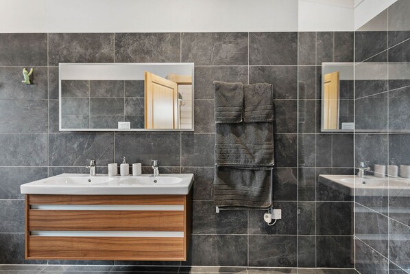 Ein modernes Badezimmer mit zwei Waschbecken, einem Holz-Waschtisch, dunklen Fliesenwänden und einem Handtuchhalter.
