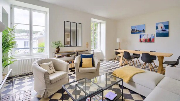 A living room with a checkered floor, a sofa, a coffee table, a dining table with chairs, and a large window with a view of buildings. - Charming town house completely renovated (Quimper)