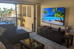 Een woonkamer met een grote flatscreentelevisie, een bank, een salontafel en uitzicht op een zwembad en palmbomen.
