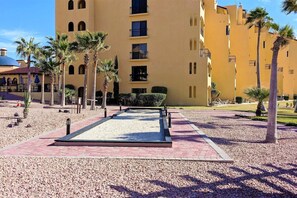 A courtyard with a central pathway, palm trees, and a yellow building with multiple balconies. - Ocean Front Resort 2-Bedroom Condo at Princesa de Peñasco with Amenities (Puerto PeÃ±asco)