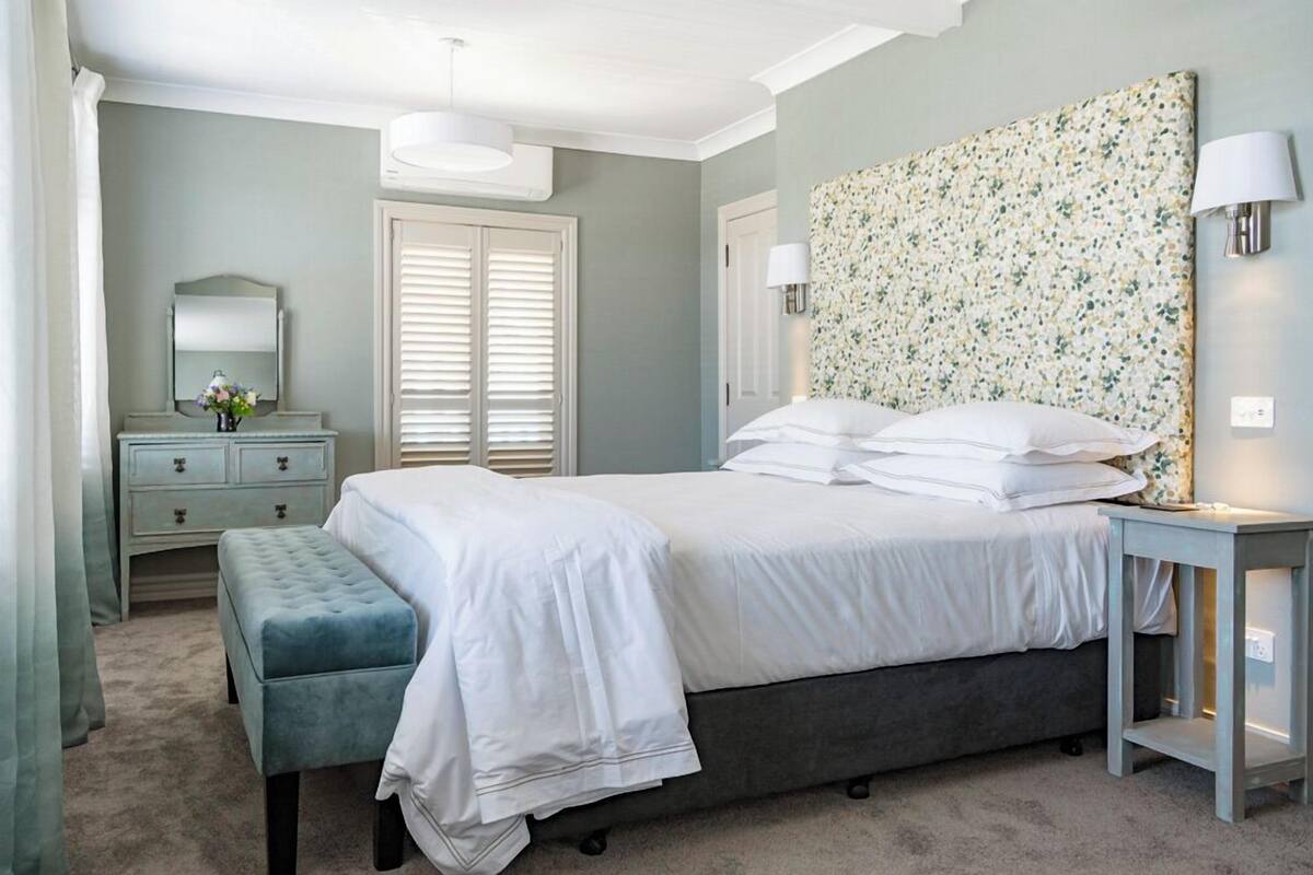 Quarto com cama, um banquinho, cômoda, espelho e luminária na parede.