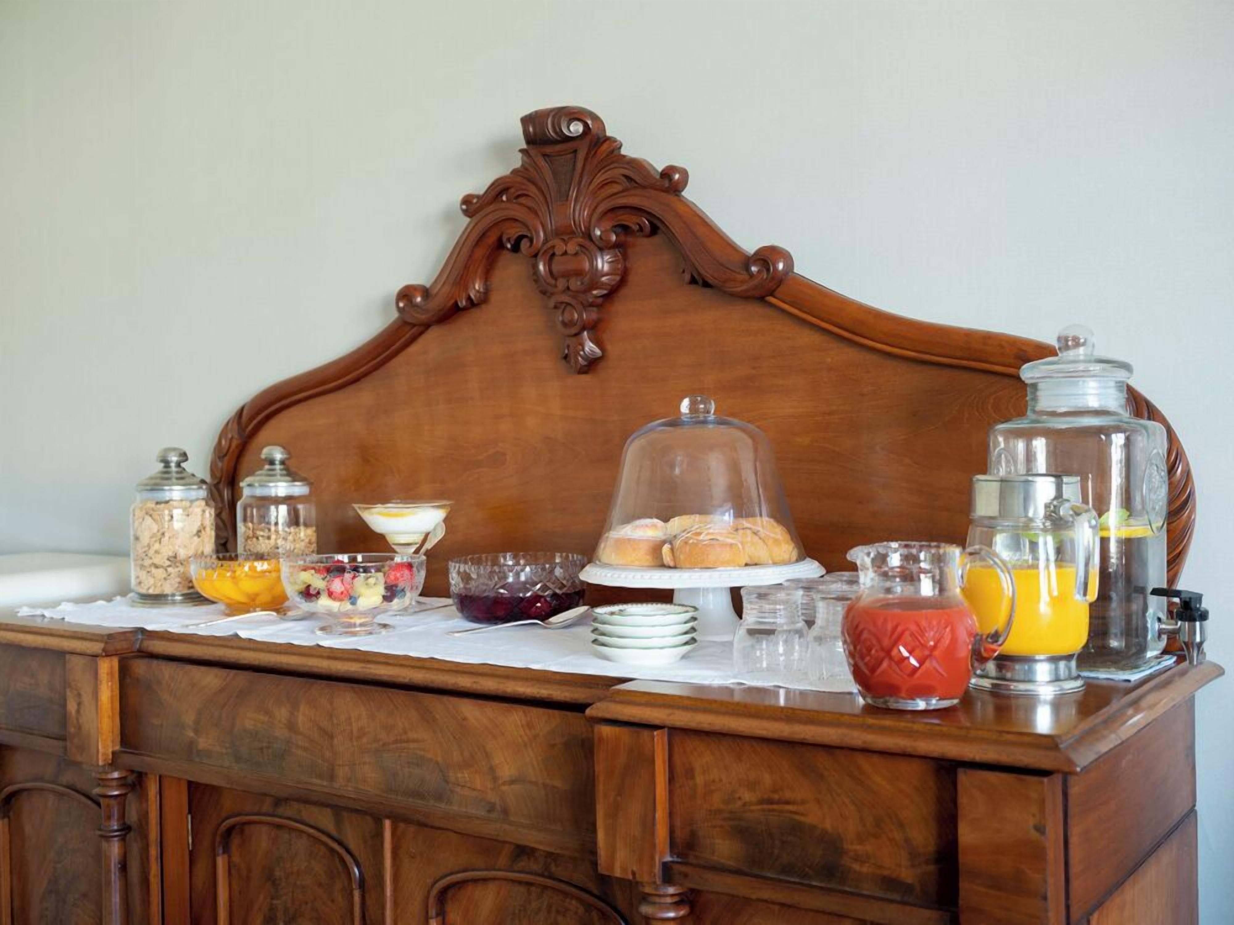 A mesa de madeira exibe um buffet de café da manhã com diversos alimentos e bebidas, em frente a uma cabeceira de cama.