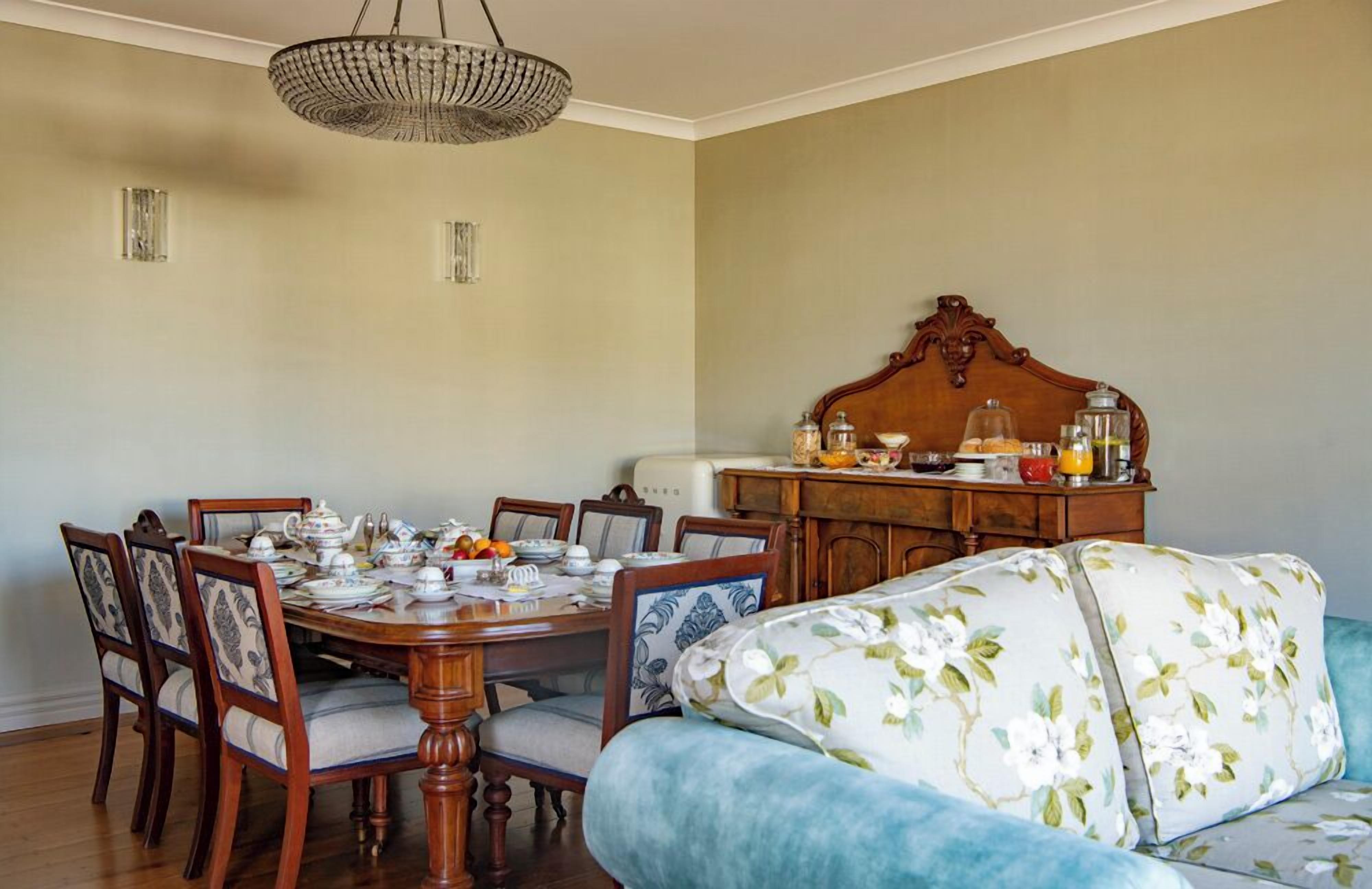 A dining area with a wooden table set for six, surrounded by upholstered chairs with floral patterns. A vintage-style chandelier hangs above, and a wooden cabinet with glass jars and a fruit bowl is to the right.