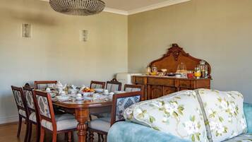 A dining area with a wooden table set for six, surrounded by upholstered chairs with floral patterns. A vintage-style chandelier hangs above, and a wooden cabinet with glass jars and a fruit bowl is to the right.