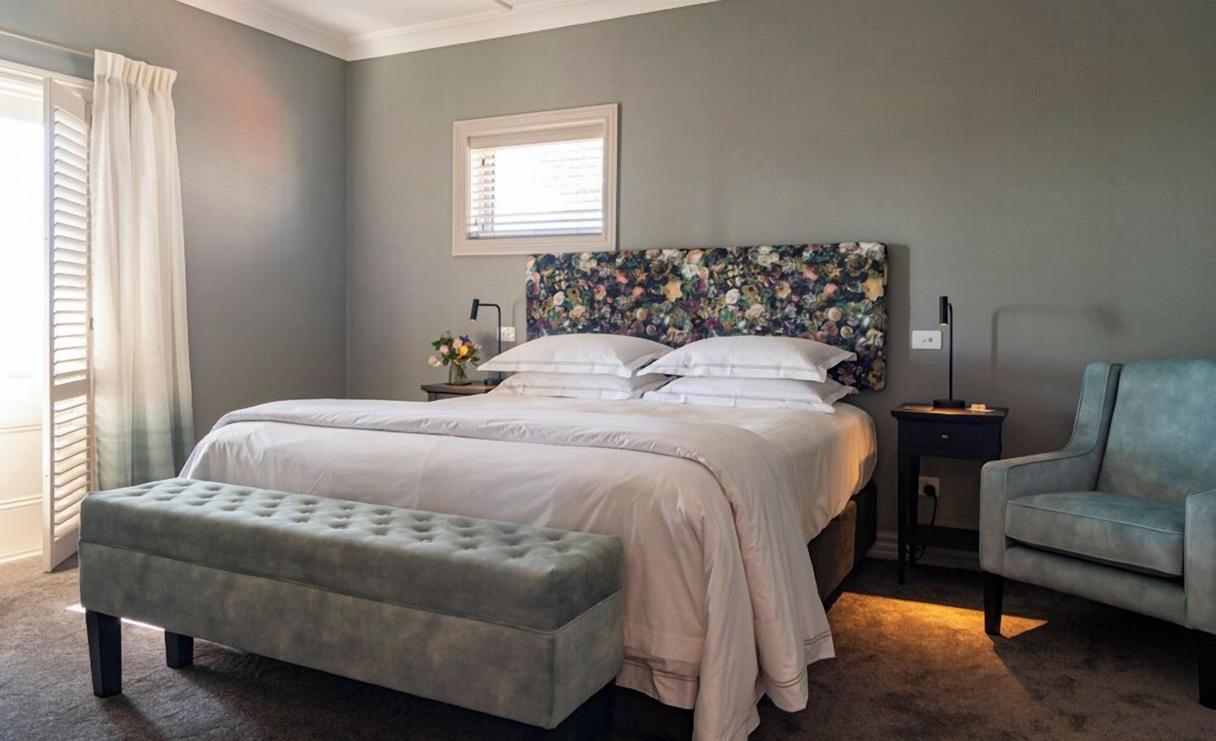 Een slaapkamer met een groot bed, een bankje, een stoel, een nachtkastje met lamp en een bloemen hoofdbord.