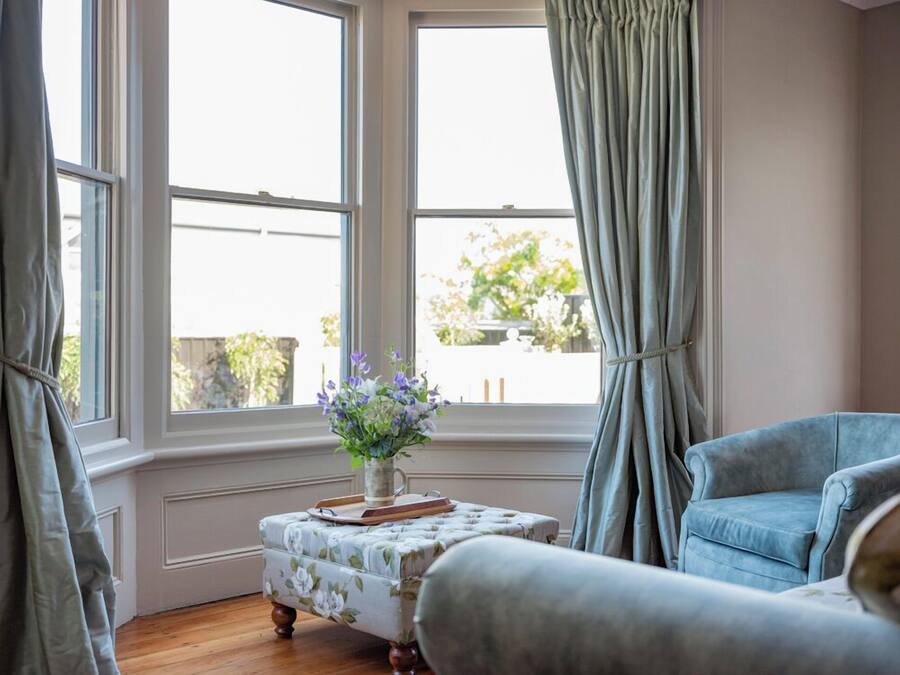 Une pièce avec une grande fenêtre, un pouf à motifs floraux, un fauteuil bleu et un vase rempli de fleurs.