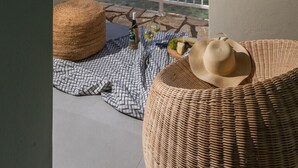 Un balcón con vista al mar, una silla de mimbre, un sombrero y una botella de vino sobre un mantel a cuadros.