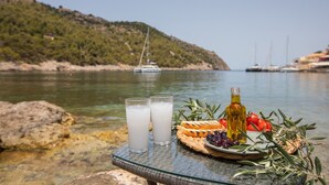Una mesa con vasos de una bebida blanca, una botella de aceite de oliva, un plato con aceitunas, tomates y romero, junto a un cuerpo de agua con barcos al fondo.