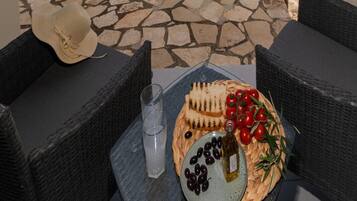 Une terrasse avec un sol en pierre, des meubles en osier, un chapeau, une assiette d’olives et de tomates, une bouteille, et une vue sur la mer et un pont.