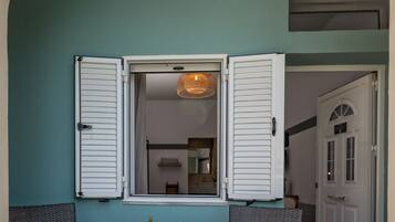 Un espacio exterior con dos sillas de mimbre, una mesa pequeña con una cesta de fruta y una puerta a la derecha.