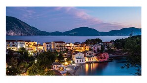 Un pueblo costero al atardecer con edificios a lo largo del paseo marítimo, un mar tranquilo y montañas al fondo.
