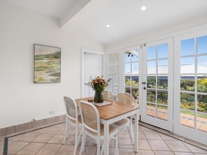 A dining area with a wooden table and white wicker chairs, a vase with flowers, and a view of a garden through large glass doors. - Roma in the Heart Mornington (Mornington)