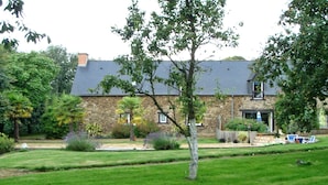 A stone house with a sloped roof, surrounded by a well-maintained lawn and various plants. - Les Cavaliers, character gîte with swimming pool, 20 minutes from the seaside (La Poterie, Lamballe)