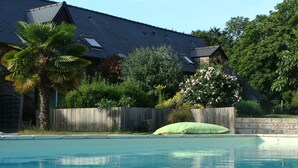 A swimming pool with a wooden fence, a palm tree, and a house with a sloped roof. - Les Cavaliers, character gîte with swimming pool, 20 minutes from the seaside (La Poterie, Lamballe)