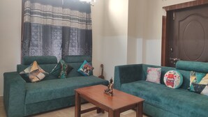 A living room with two green sofas, a wooden coffee table, and patterned curtains. - Homocation The Corporates Homestay (Guwahati)