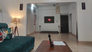 A spacious living room with a sofa, a small table with a Buddha statue, a ceiling fan, and a neon sign on the wall. - Homocation The Corporates Homestay (Guwahati)