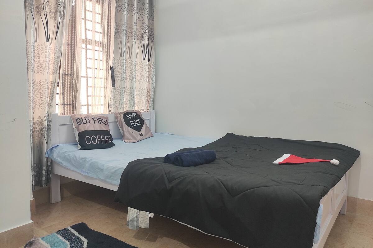 A single bed with a black bedspread, two pillows with text, a blue blanket, a red and white hat, and a patterned rug.