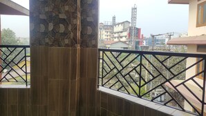 A balcony with a stone wall and metal railing, overlooking a cityscape with buildings and a tower. - Homocation The Corporates Homestay (Guwahati)