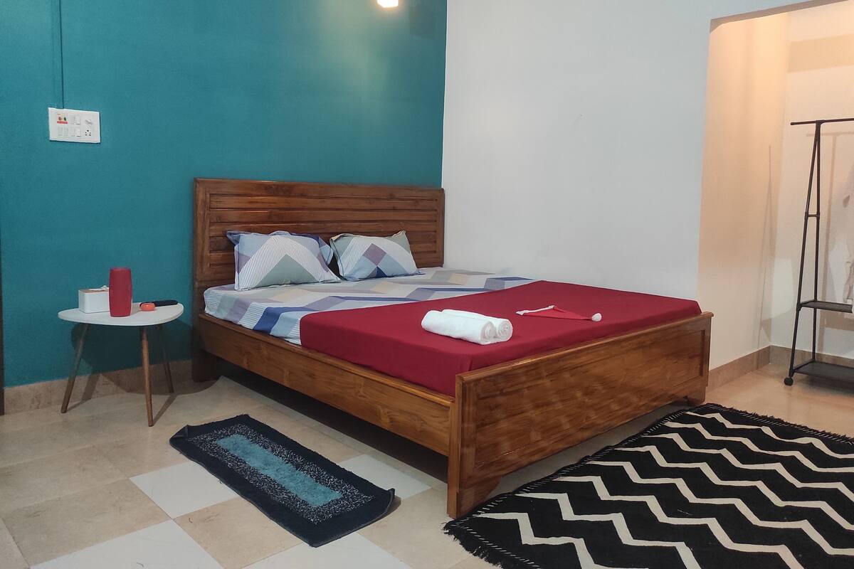 A neatly arranged bedroom with a wooden bed, a small bedside table with a red object, and a patterned rug on the floor.