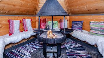 A cozy cabin interior with a central wood-burning stove, plush seating with patterned cushions, and a wooden ceiling.