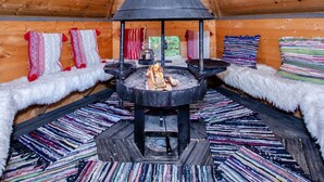 A cozy cabin interior with a central wood-burning stove, plush seating with colorful blankets, and a wooden ceiling.