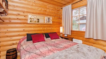 Ein Schlafzimmer in einer Blockhütte mit einem Bett, einem Nachttisch, einem Fenster mit Vorhängen und einer Holzbalkendecke.