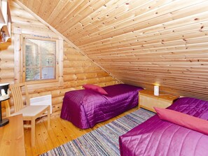 Une cabane chaleureuse avec un plafond en pente, un lit avec une literie violette, un petit bureau et une fenêtre avec des stores.