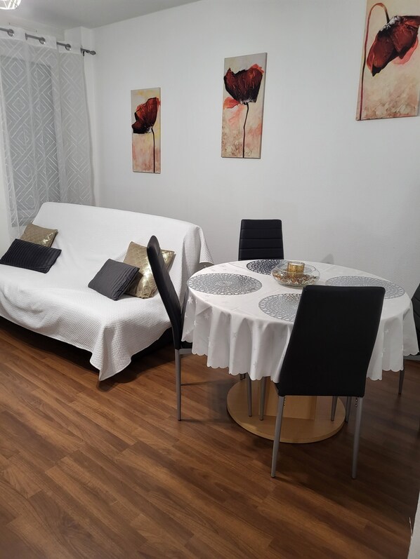 A dining area with a round table, black chairs, and a white tablecloth. There is a white sofa with patterned cushions in the background.