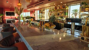 A bar area with a marble countertop, bar stools, a fruit bowl, and a variety of bottles and glasses. - The Eden (Santa Monica)