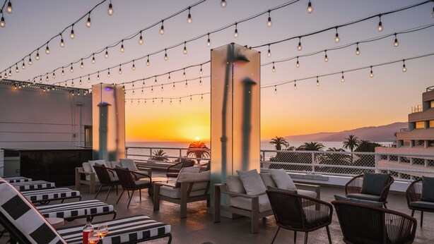 A rooftop lounge with lounge chairs and tables, string lights, and a sunset view. - The Eden (Santa Monica)