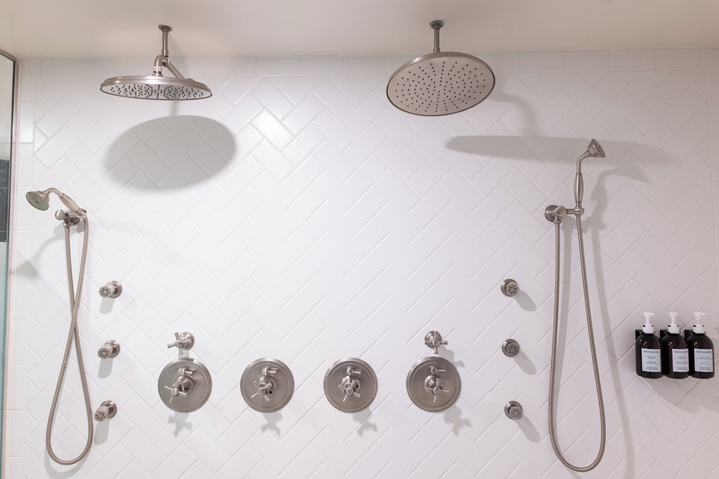 Un baño con pared de azulejos blancos en patrón de espiga, un cabezal de ducha, una ducha de mano, un grifo empotrado y tres botellas de champú.