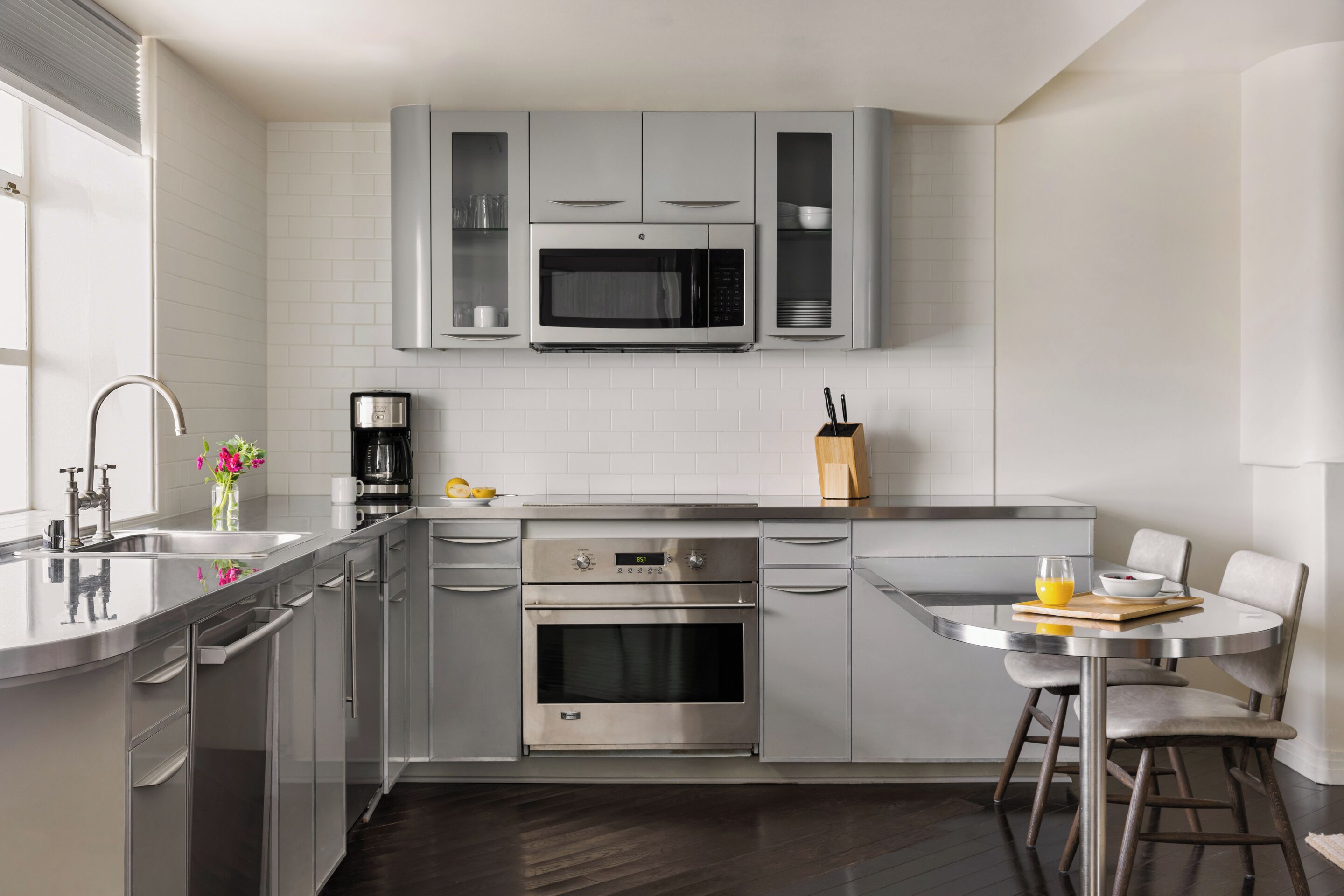 A modern kitchen with stainless steel appliances, a dining area with a round table and bar stools, and a large window providing natural light.