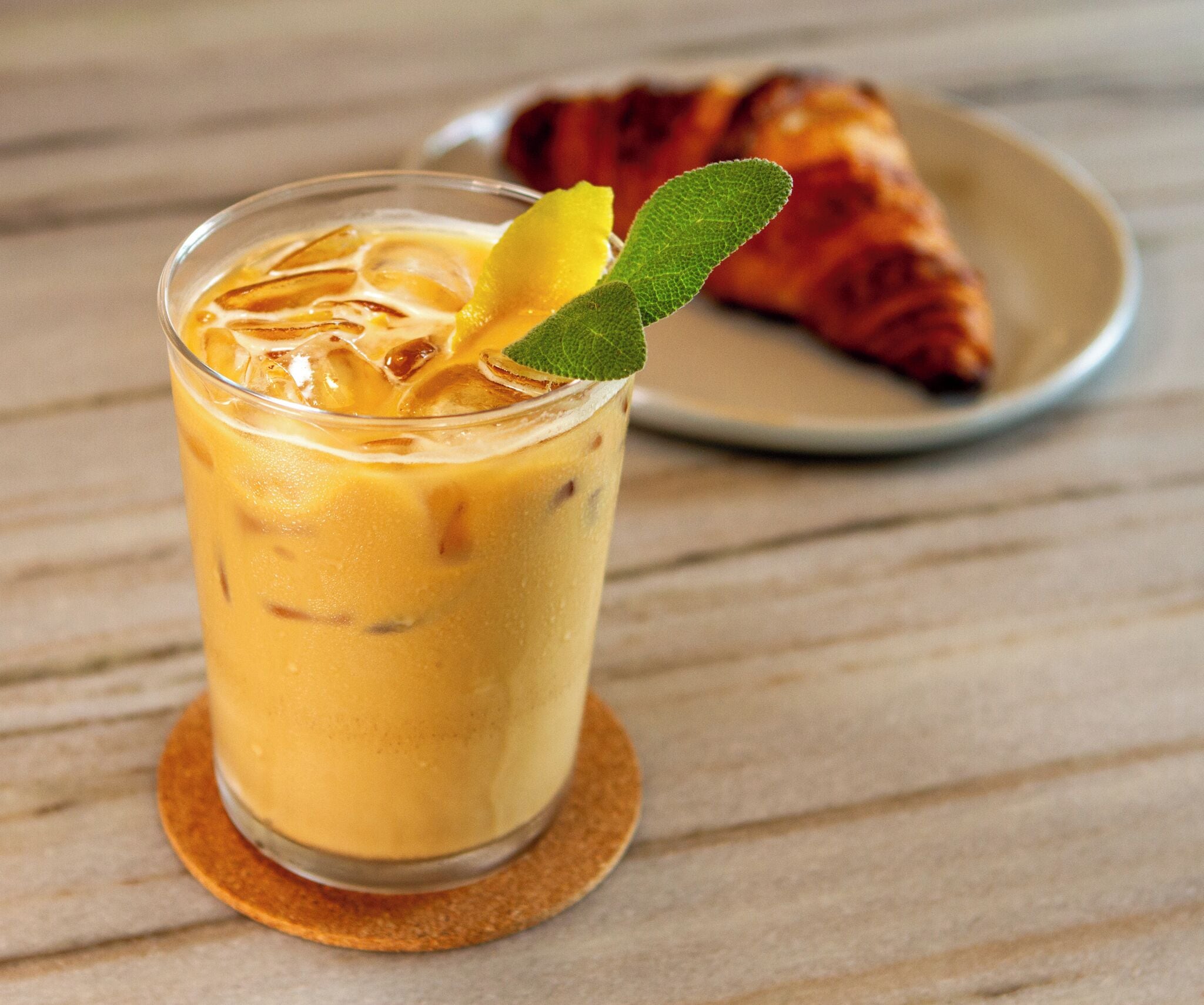A glass of iced coffee with a lemon slice and mint on the rim, placed on a coaster. In the background, there is a croissant on a plate.