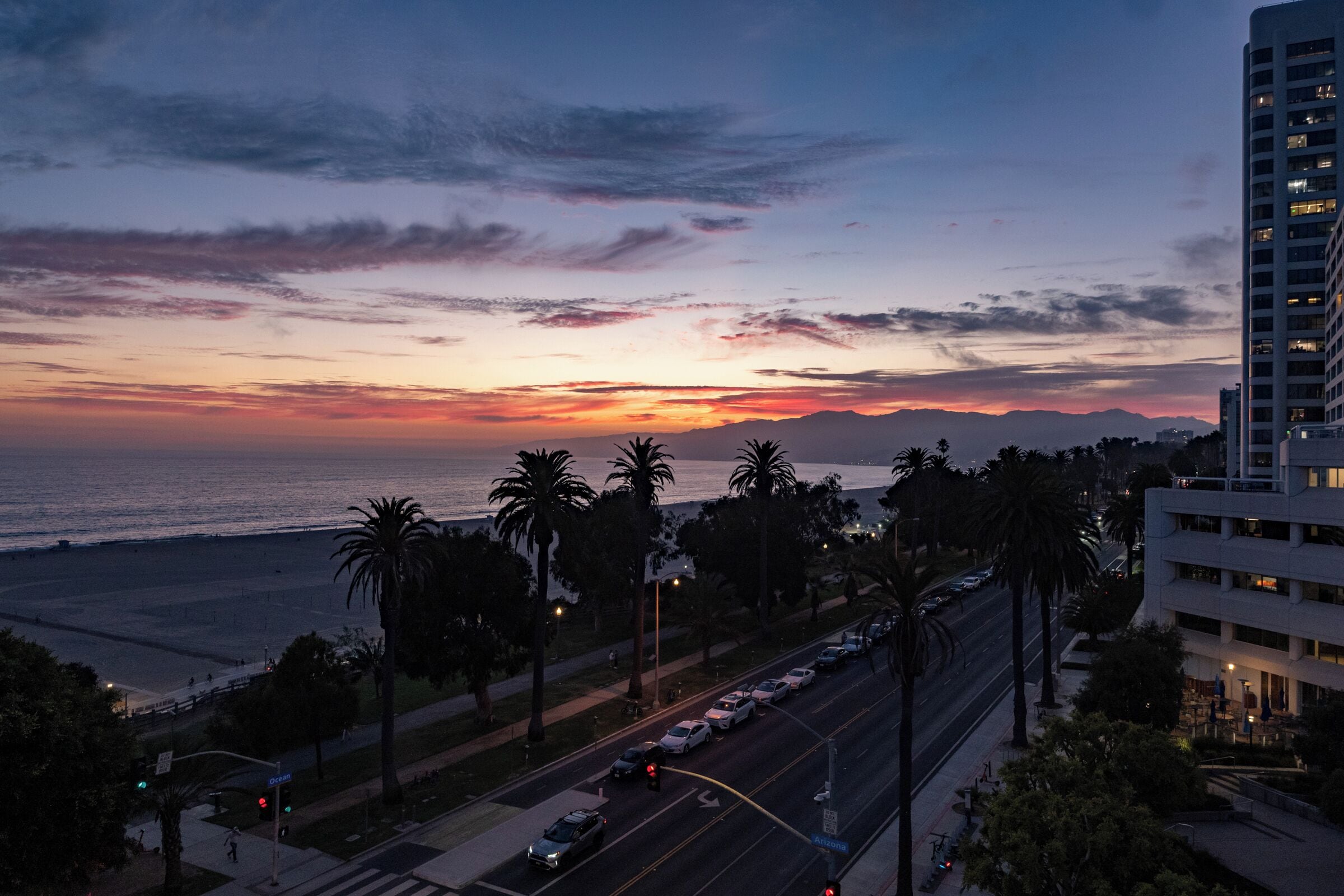 A coastal cityscape at dusk with a sunset, palm trees, and a multi-story building.